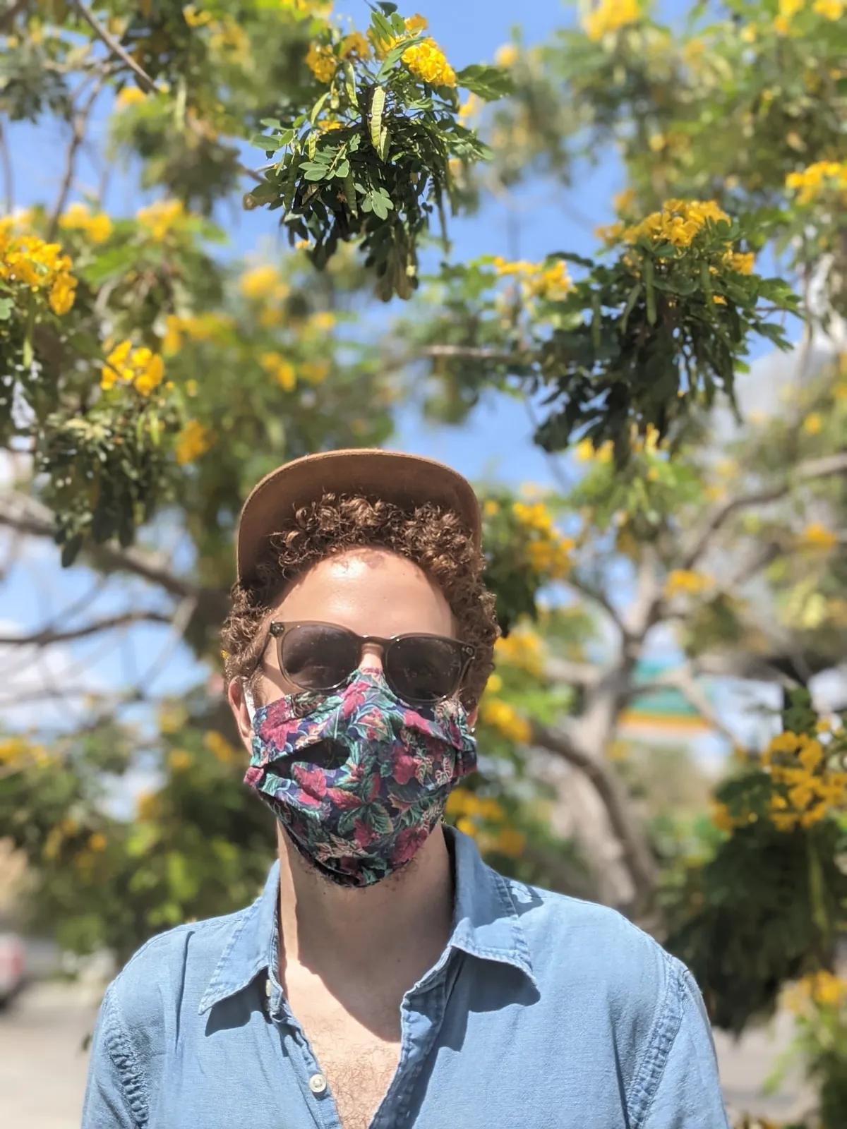 David in a brown hat, sunglasses and a floral face mask, under a tree of yellow blossom.