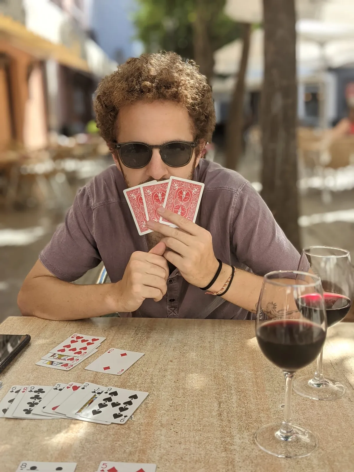 David playing cards at an outdoor table with a glass of red wine, holding his hand up to his face — still in sunglasses.